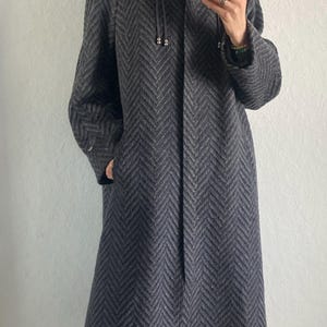 May include: A long, dark gray herringbone pattern coat with a high collar and a hood with drawstrings. The coat has pockets and is worn with gray socks and black and white sneakers.