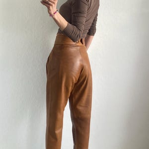 May include: A brown long-sleeve top and high-waisted leather pants. The pants are a light brown color with a wide waistband. The person is wearing black and white sneakers with a leopard print design.