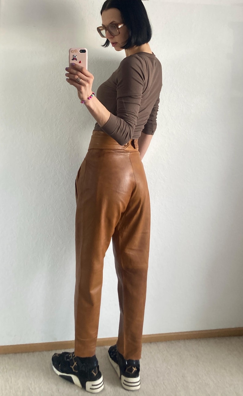 May include: A brown long-sleeve top and high-waisted leather pants. The pants are a light brown color with a wide waistband. The person is wearing black and white sneakers with a leopard print design.