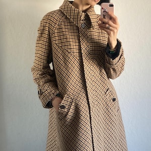 Può includere: Un cappotto in stile vintage con motivo a quadri marrone e nero. Il cappotto presenta un colletto classico, chiusure con bottoni e tasche. Il cappotto è lungo fino al ginocchio e ha maniche lunghe.