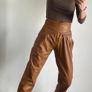 May include: A person wearing a brown long-sleeve shirt and high-waisted brown leather pants. The person is also wearing black and tan sneakers. The outfit is captured in a full-length shot.