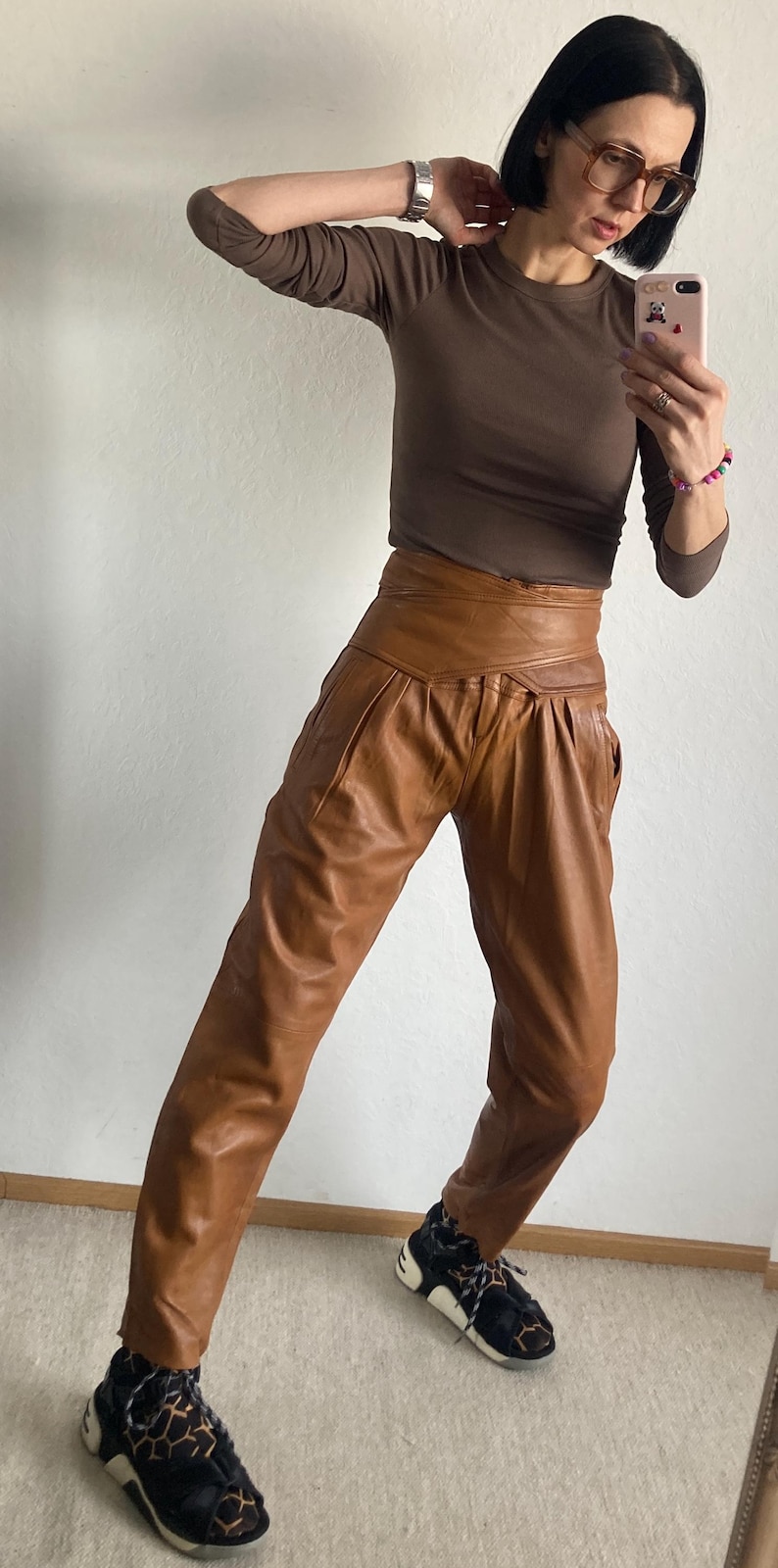 May include: A person wearing a brown long-sleeve shirt and high-waisted brown leather pants. The person is also wearing black and tan sneakers. The outfit is captured in a full-length shot.