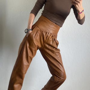 May include: A woman wearing a brown long-sleeve shirt and high-waisted, pleated brown leather pants. She is also wearing black and white sneakers with a geometric pattern. A watch is visible on her left wrist.