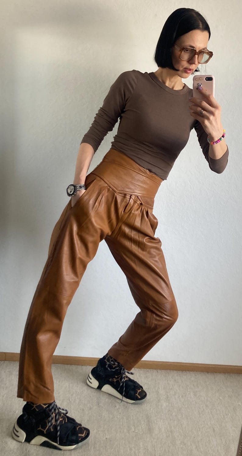 May include: A woman wearing a brown long-sleeve shirt and high-waisted, pleated brown leather pants. She is also wearing black and white sneakers with a geometric pattern. A watch is visible on her left wrist.