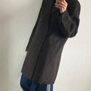 May include: A dark brown double-breasted coat with large buttons and a notched collar. The coat is paired with dark blue jeans with white stripes down the side and red sneakers.