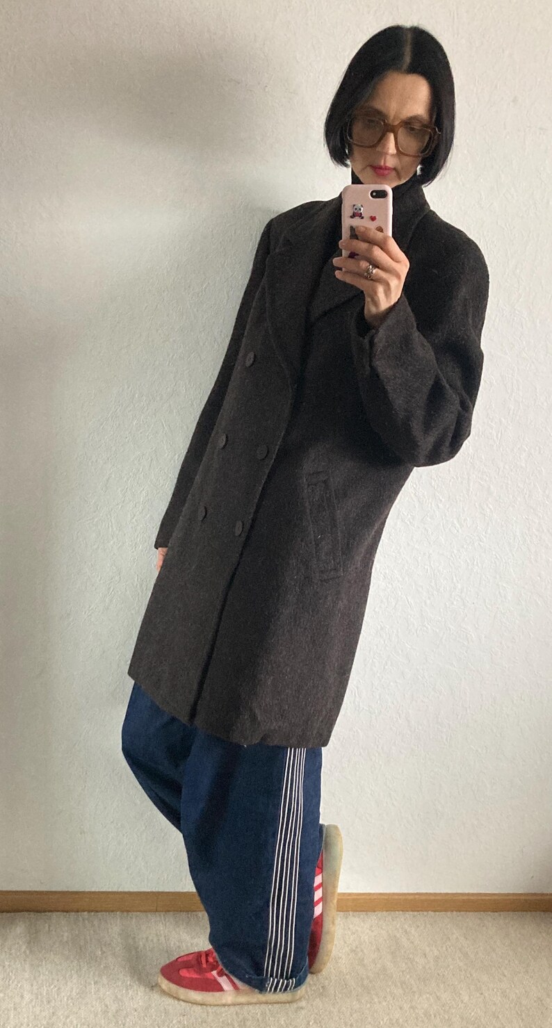 May include: A dark brown double-breasted coat with large buttons and a notched collar. The coat is paired with dark blue jeans with white stripes down the side and red sneakers.