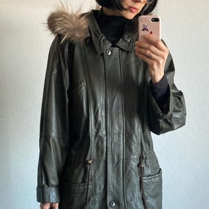 May include: Olive green leather jacket with a fur-trimmed hood. The jacket features a button-down front, pockets, and drawstring details. A classic outerwear piece for colder weather.