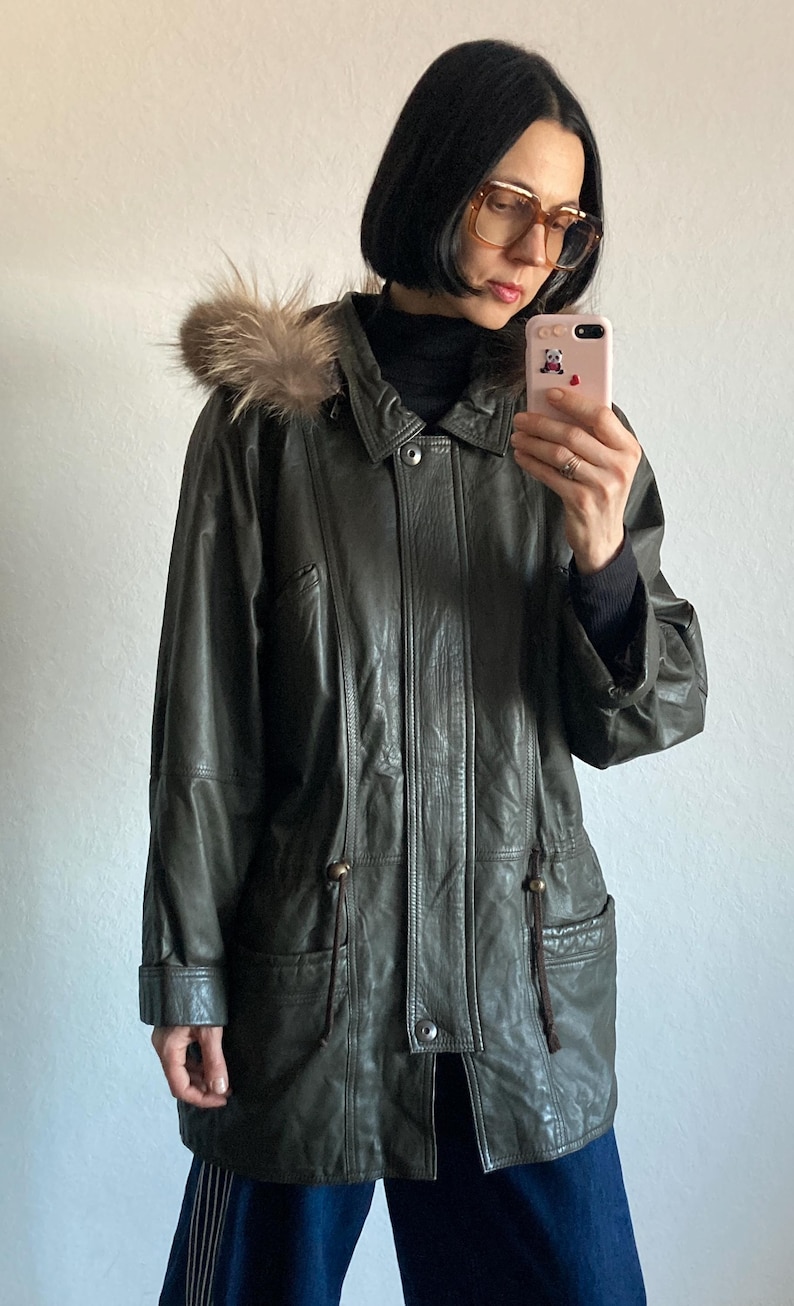 May include: Olive green leather jacket with a fur-trimmed hood. The jacket features a button-down front, pockets, and drawstring details. A classic outerwear piece for colder weather.