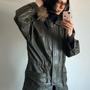May include: Olive green leather jacket with a fur-trimmed hood. The jacket has a button-down front, pockets, and a drawstring waist. A stylish outerwear piece for colder weather.