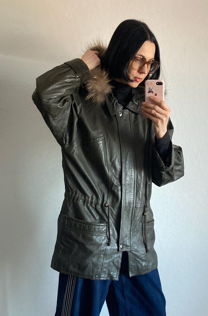 May include: Olive green leather jacket with a fur-trimmed hood. The jacket has a button-down front, pockets, and a drawstring waist. A stylish outerwear piece for colder weather.