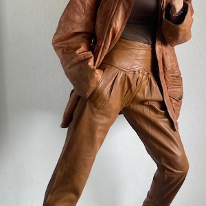 May include: A brown leather jacket and matching high-waisted leather pants. The jacket has a collared neckline and pockets. The pants have a pleated front. The outfit is paired with black and tan sneakers.