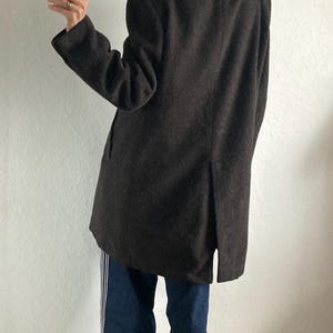 May include: A dark brown coat with a high collar and a split in the back. The person is wearing blue jeans with white stripes down the sides and red and white shoes.