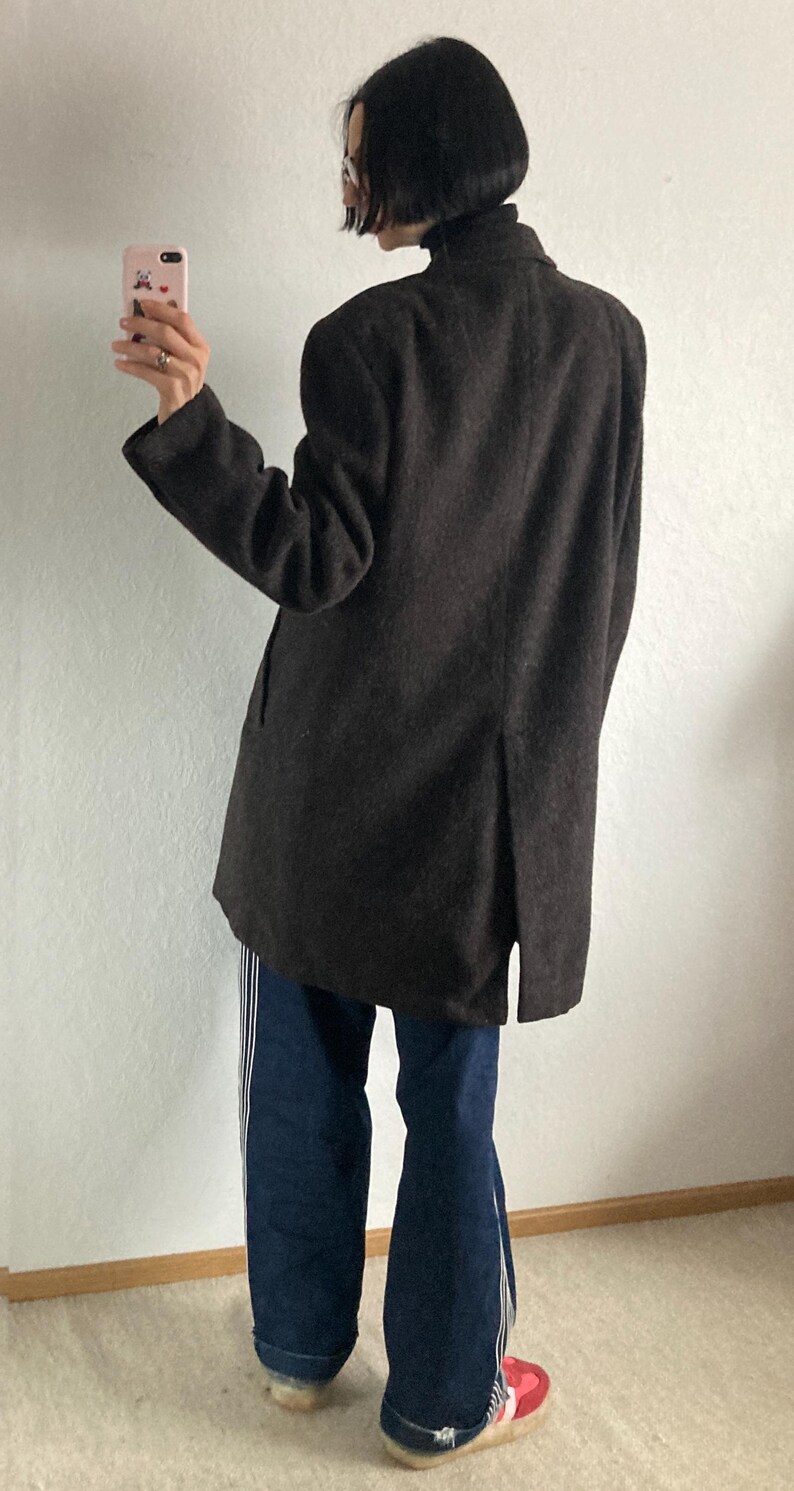 May include: A dark brown coat with a high collar and a split in the back. The person is wearing blue jeans with white stripes down the sides and red and white shoes.