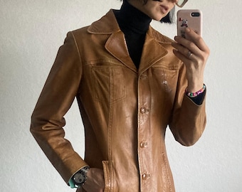 May include: A tan leather jacket with a classic collar, button closure, and front pockets. The jacket has a tailored fit and is worn over a black turtleneck. The person is wearing glasses and holding a pink phone.