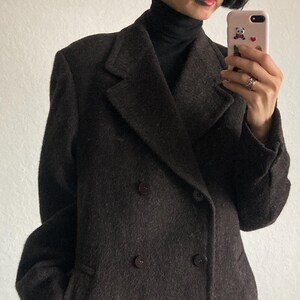 May include: A dark brown double-breasted coat with large lapels and dark buttons. The coat has a classic design and appears to be made of a textured fabric. The person wearing the coat is also wearing a black turtleneck.