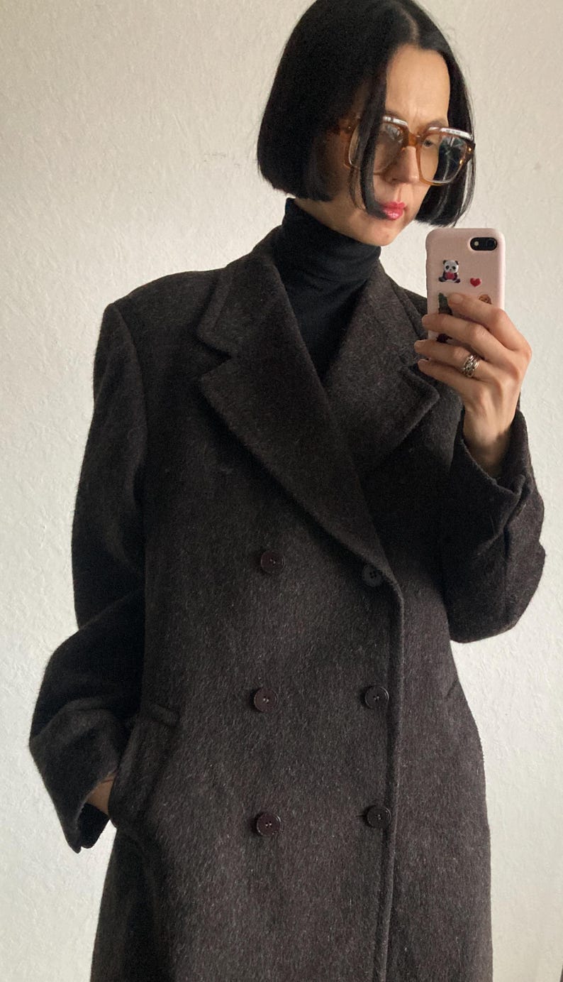 May include: A dark brown double-breasted coat with large lapels and dark buttons. The coat has a classic design and appears to be made of a textured fabric. The person wearing the coat is also wearing a black turtleneck.