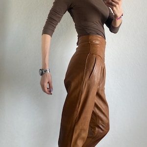 May include: A brown long-sleeved top and high-waisted, wide-leg leather pants. The pants are a lighter shade of brown. The person is wearing black and white sneakers. The outfit is styled with a silver watch and glasses.