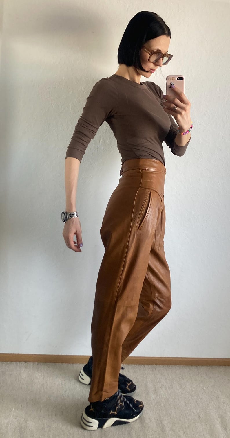 May include: A brown long-sleeved top and high-waisted, wide-leg leather pants. The pants are a lighter shade of brown. The person is wearing black and white sneakers. The outfit is styled with a silver watch and glasses.