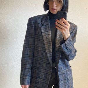 Könnte beinhalten: Ein grauer und blauer karierter Blazer mit einem schwarzen Rollkragenpullover und einer schwarzen Jeans. Der Blazer hat einen einzigen Knopfverschluss und zwei Taschen.