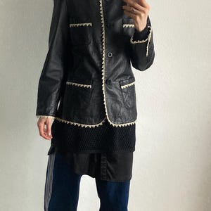 May include: Black leather jacket with white trim and a single button closure. The jacket is paired with dark blue pants with white stripes and yellow sneakers. The outfit is a mix of vintage and modern styles.