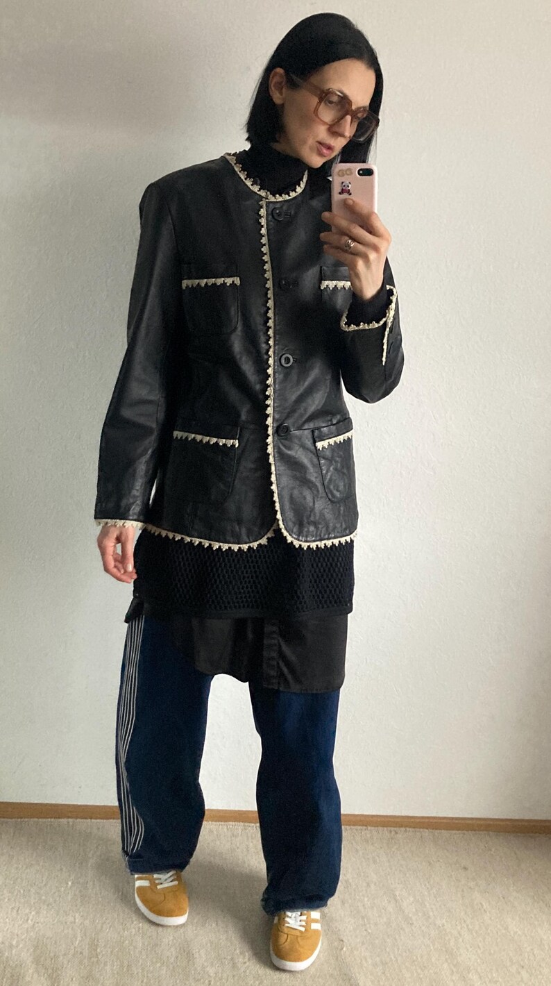 May include: Black leather jacket with white trim and a single button closure. The jacket is paired with dark blue pants with white stripes and yellow sneakers. The outfit is a mix of vintage and modern styles.