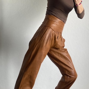 May include: A brown long-sleeve top and high-waisted brown leather pants. The pants have a pleated front and a wide waistband. The person is wearing black and white sneakers with a patterned design.