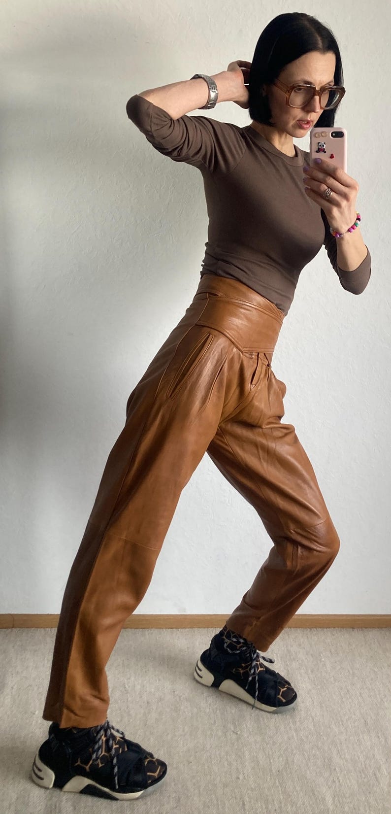 May include: A brown long-sleeve top and high-waisted brown leather pants. The pants have a pleated front and a wide waistband. The person is wearing black and white sneakers with a patterned design.