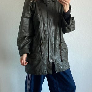 May include: Dark green leather jacket with a fur-trimmed hood. The jacket has a button-down front, pockets, and drawstring details. The person is wearing dark blue pants with white stripes and gray shoes.