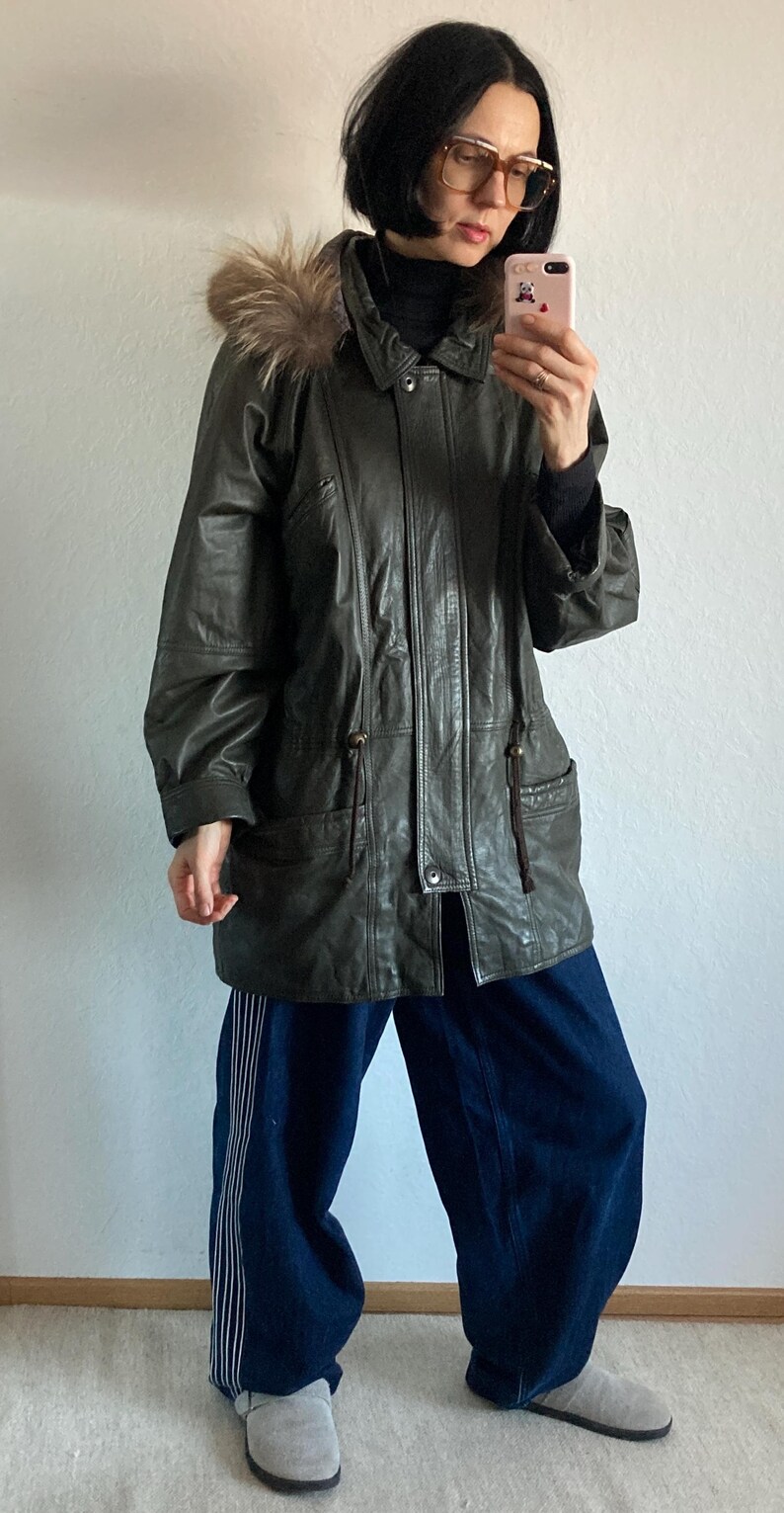 May include: Dark green leather jacket with a fur-trimmed hood. The jacket has a button-down front, pockets, and drawstring details. The person is wearing dark blue pants with white stripes and gray shoes.