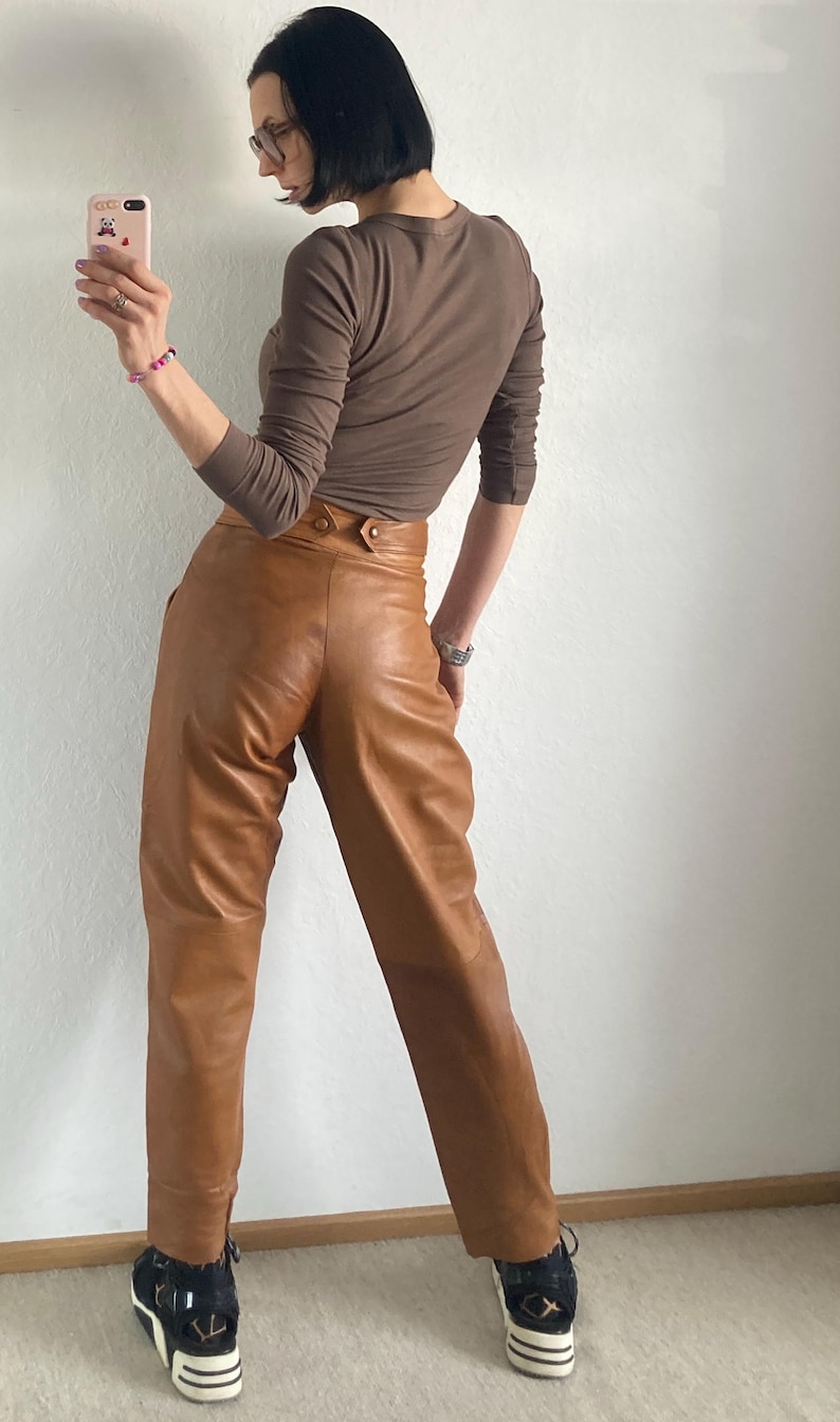 May include: A brown long-sleeve shirt and tan leather pants. The pants have a high waist and button details. The person is wearing black and white sneakers.