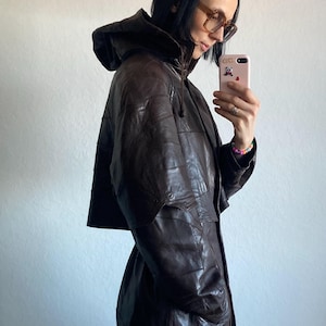 May include: A dark brown leather jacket with a hood. The jacket has a patchwork design and a drawstring. It features pockets and a zipper closure. The person in the image is wearing glasses and holding a pink phone.