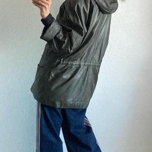 May include: An olive green hooded parka with a fur-trimmed hood. The parka is worn with dark blue jeans with white stripes down the sides. The person is holding a pink phone.