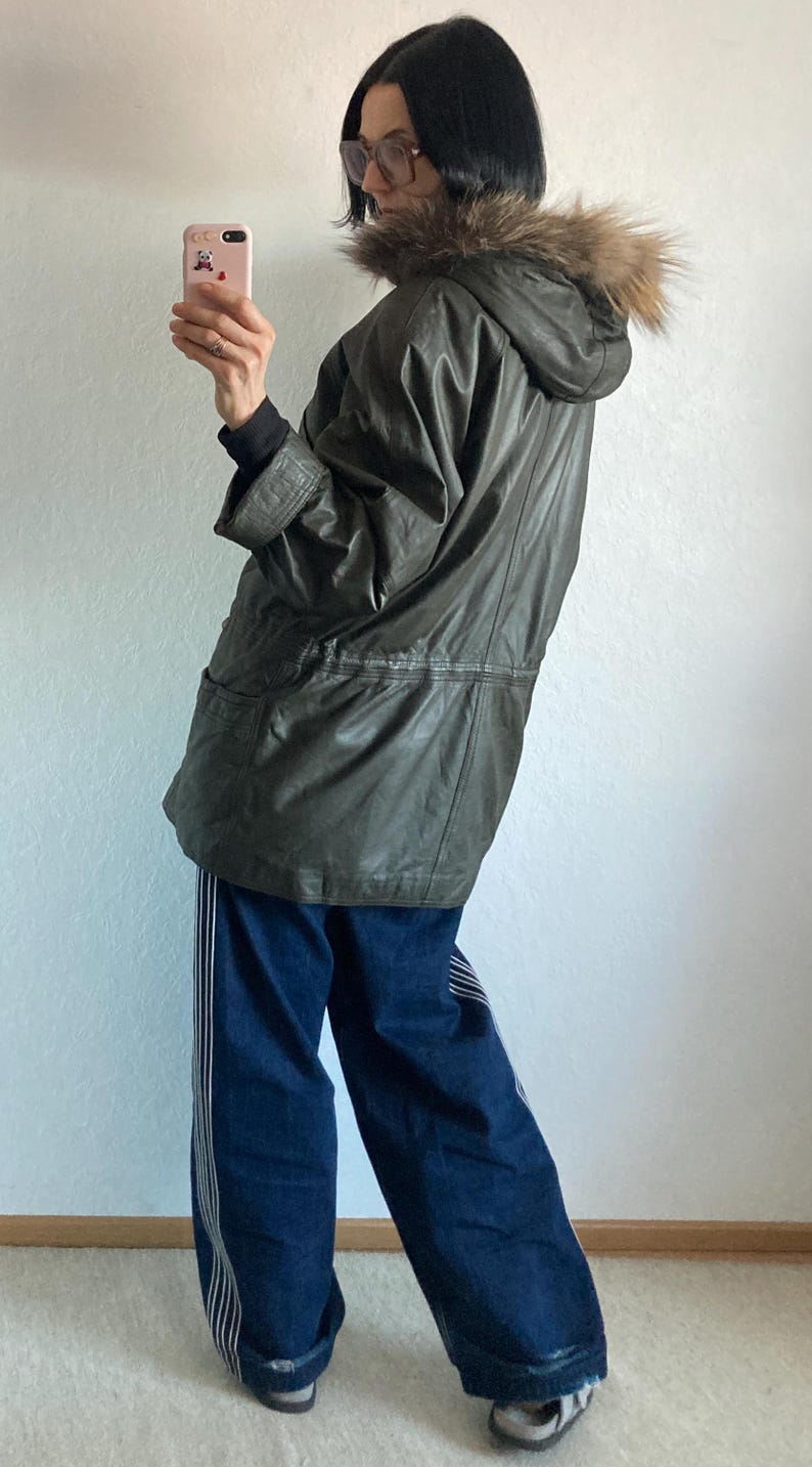 May include: An olive green hooded parka with a fur-trimmed hood. The parka is worn with dark blue jeans with white stripes down the sides. The person is holding a pink phone.