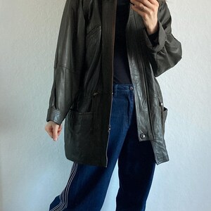 May include: Dark green leather jacket with a fur-trimmed hood. The jacket has a button-up front and pockets. The person is wearing a black turtleneck, dark blue jeans with white stripes, and gray shoes.