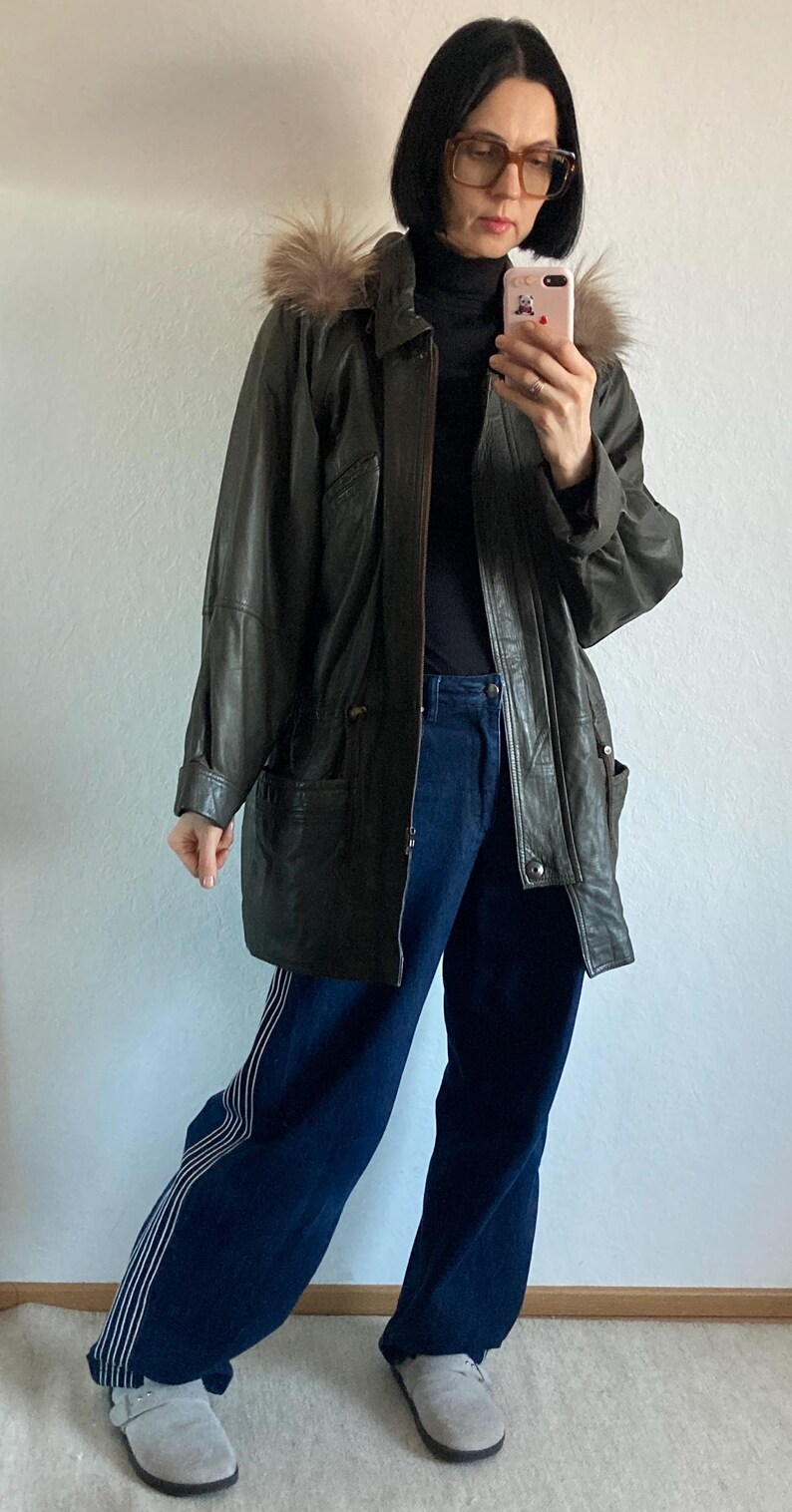 May include: Dark green leather jacket with a fur-trimmed hood. The jacket has a button-up front and pockets. The person is wearing a black turtleneck, dark blue jeans with white stripes, and gray shoes.