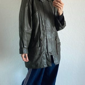 May include: A dark green leather jacket with a fur-trimmed hood. The jacket has a button-up front, pockets, and a drawstring waist. The person is wearing dark blue pants and gray clogs.
