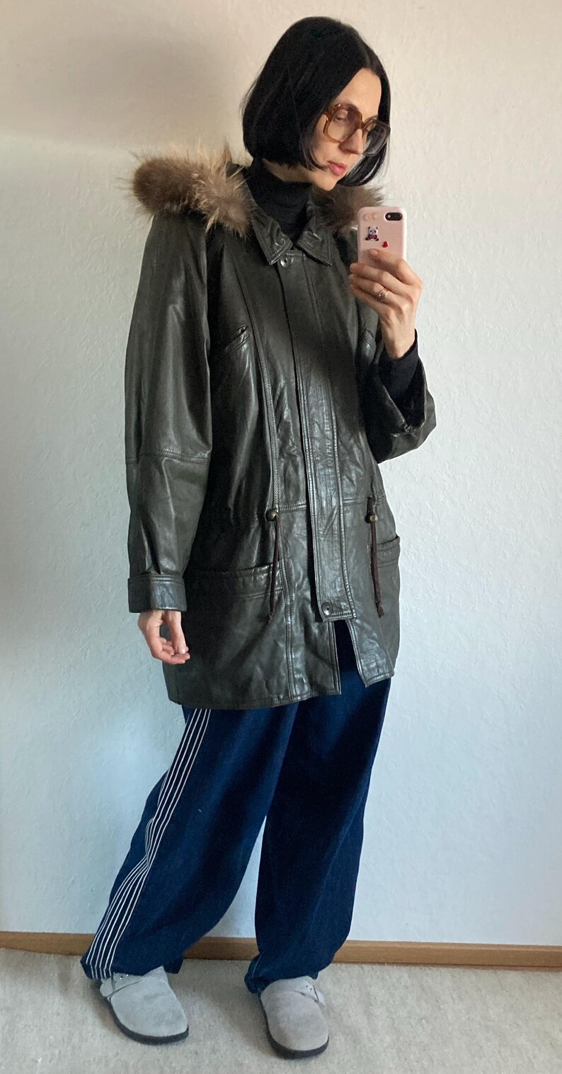 May include: A dark green leather jacket with a fur-trimmed hood. The jacket has a button-up front, pockets, and a drawstring waist. The person is wearing dark blue pants and gray clogs.