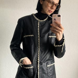 May include: Black leather jacket with cream-colored lace trim around the collar, pockets, and cuffs. The jacket has a button-up front and two front pockets. A classic, vintage-inspired outerwear piece.