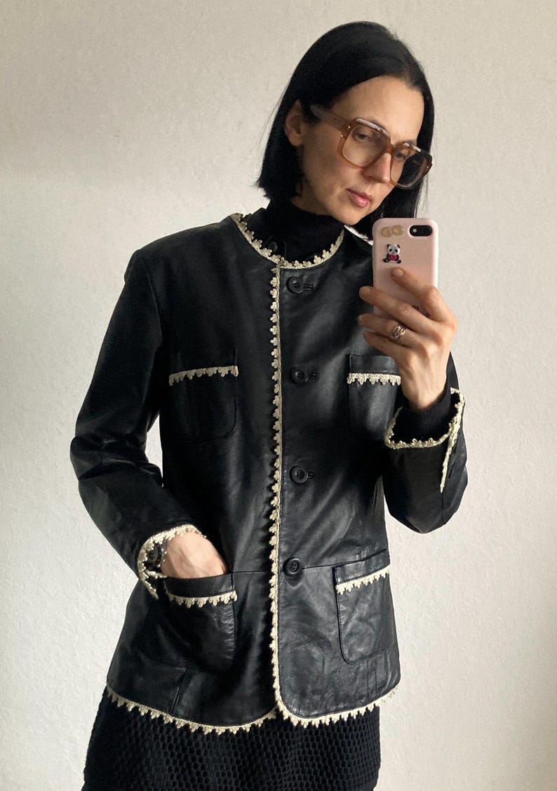 May include: Black leather jacket with cream-colored lace trim around the collar, pockets, and cuffs. The jacket has a button-up front and two front pockets. A classic, vintage-inspired outerwear piece.