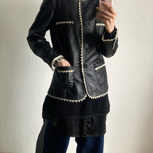 May include: Black leather jacket with white trim and pockets. The jacket has a button-up front and is worn with a black top, dark blue pants with white stripes, and yellow sneakers.