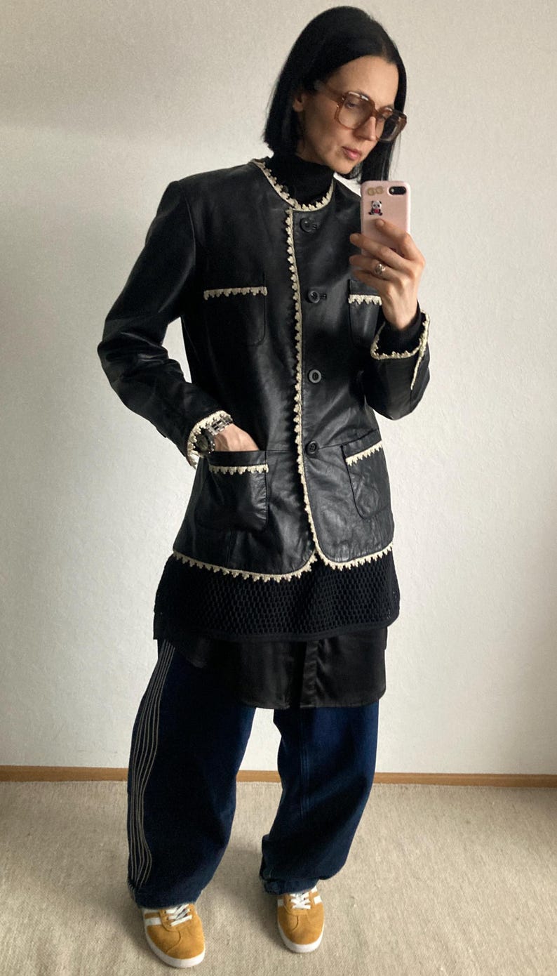 May include: Black leather jacket with white trim and pockets. The jacket has a button-up front and is worn with a black top, dark blue pants with white stripes, and yellow sneakers.