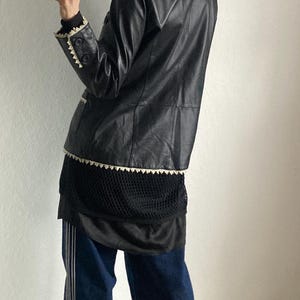 May include: A black leather jacket with white trim is worn over a black mesh top and a black skirt. The outfit is paired with dark blue jeans with white stripes down the sides.