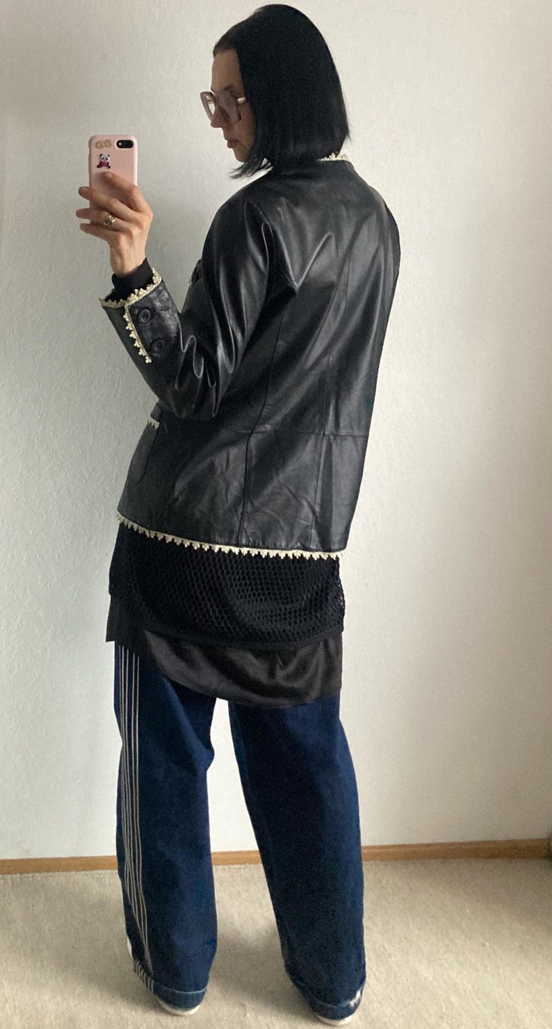 May include: A black leather jacket with white trim is worn over a black mesh top and a black skirt. The outfit is paired with dark blue jeans with white stripes down the sides.