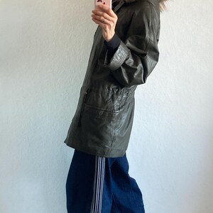 May include: An olive green leather jacket with a fur-trimmed hood. The jacket is paired with dark blue jeans with white stripes down the side. The person is holding a pink phone.
