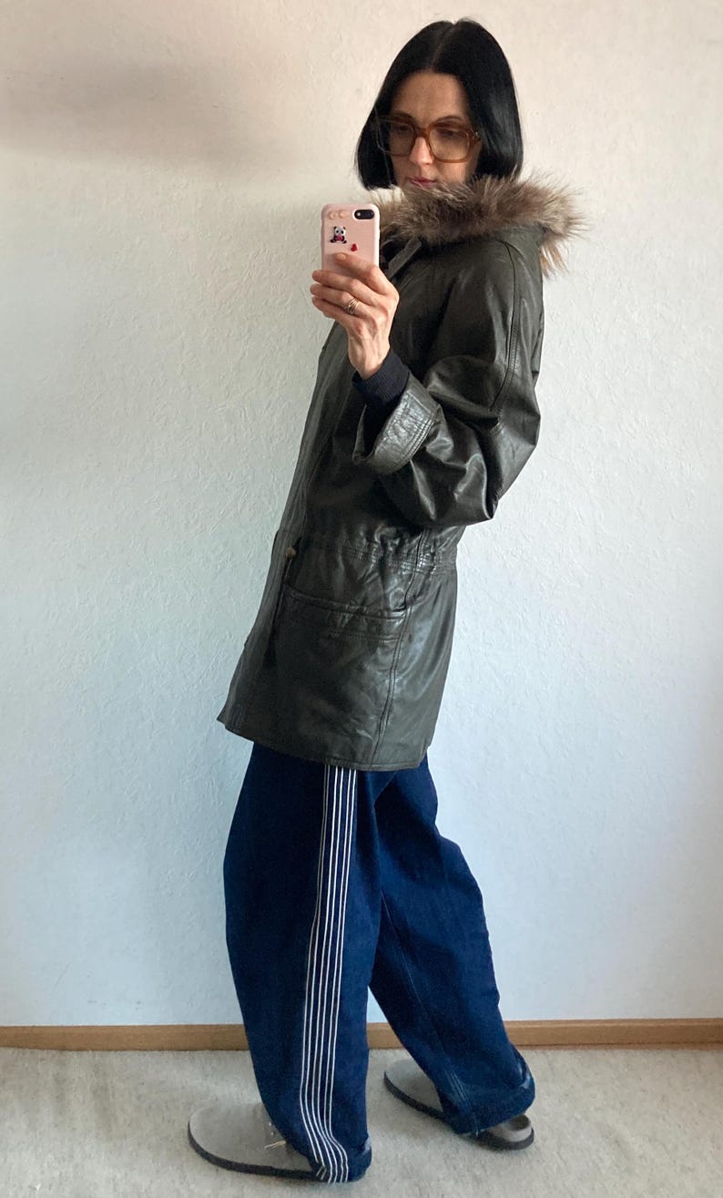 May include: An olive green leather jacket with a fur-trimmed hood. The jacket is paired with dark blue jeans with white stripes down the side. The person is holding a pink phone.