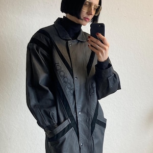 Può includere: Una giacca di pelle nera con un design geometrico e un colletto nero. La giacca ha una chiusura con bottoni e tasche sui lati.