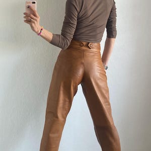 May include: A person wearing a brown long-sleeve shirt and high-waisted, tan leather pants. The pants have a button closure. The person is wearing black and white platform sneakers.