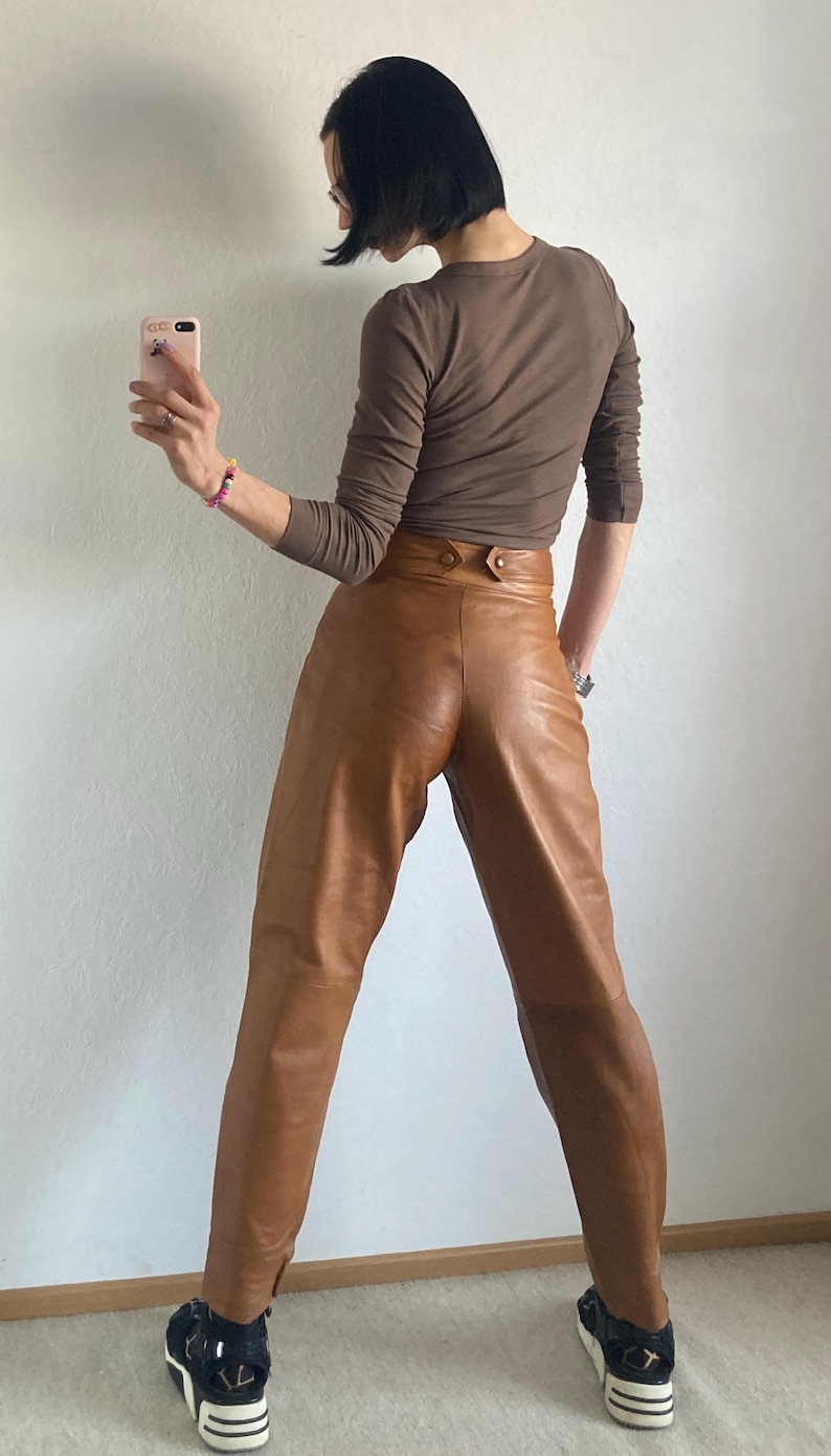 May include: A person wearing a brown long-sleeve shirt and high-waisted, tan leather pants. The pants have a button closure. The person is wearing black and white platform sneakers.