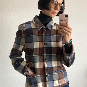 Puede incluir: Chaqueta a cuadros en tonos marrón, azul y crema. La chaqueta tiene cuello, botones y un pequeño bolsillo. La persona lleva un cuello alto negro y vaqueros azul oscuro.
