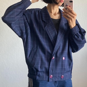 May include: A navy blue plaid blazer with pink buttons. The jacket has a double-breasted design and a gathered waist. The model is wearing the blazer with dark wash denim pants.